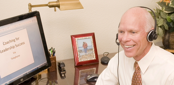 Dr. Merlin Switzer using headphones in front of the computer providing coaching over the phone with the banner, Coaching for Leadership Success, written on the screen.
